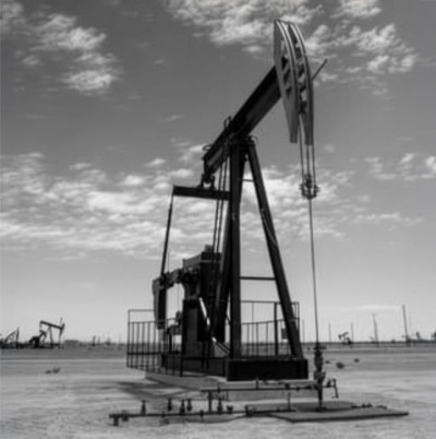 A stark, black and white photograph of an oil pumpjack, or nodding donkey, dominating the foreground in a barren, flat landscape. The machine stands tall against a bright sky filled with scattered cumulus clouds, with several other pumpjacks visible in the distance.