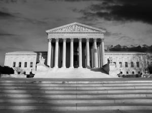 A black and white, dramatic photograph of the Supreme Court of the United States building in Washington D.C. The neoclassical structure, with its central portico and tall columns, is viewed from the base of the wide exterior steps, with dark clouds framing the top of the building.