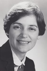 A black and white headshot of Carol Dinkins, a professional woman with short dark hair, wearing a suit jacket, a collared shirt, and a tie, smiling slightly toward the camera.