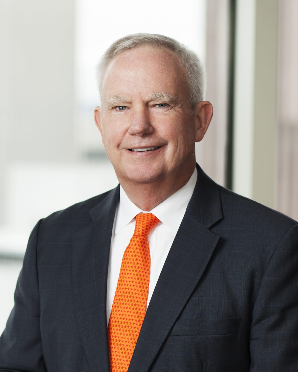 A professional headshot of Joseph C. Dilg, a man with gray hair, wearing a dark suit with a subtle plaid pattern, a white shirt, and a bright orange tie with a small pattern. He is smiling and positioned in front of a blurred window view of a modern building.
