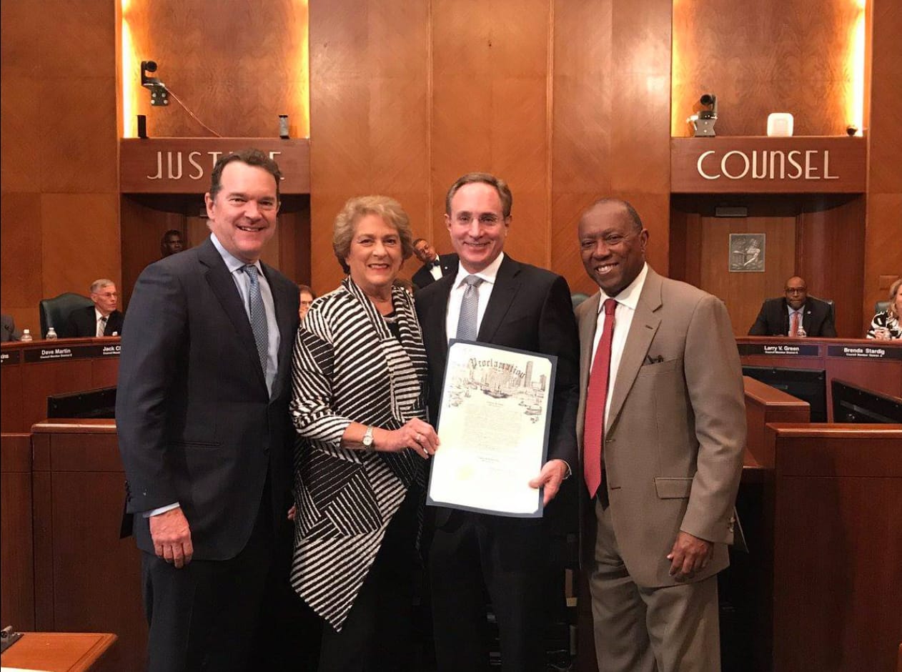 A photograph taken at the Houston City Council chambers shows four people, including two men in dark suits and a woman in a black and white striped outfit, posing with an official proclamation document. From left to right: an attorney, Council Member Ellen Cohen, another attorney holding the proclamation, and Mayor Sylvester Turner in a light brown suit and red tie. The background features wood paneling and signs reading "JUSTICE" and "COUNSEL."