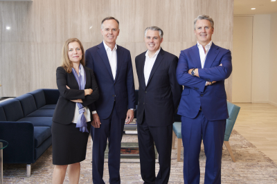 A professional group portrait of the four-member Executive Committee in a modern, well-lit office lounge. From left to right: Hilary Preston, Keith Fullenweider, Jim Fox, and Michael Holmes. They are all wearing business suits, with the three men in dark suits and the woman in a black suit with a light purple scarf. A blue sofa, an orchid, and wood-paneled walls are visible in the background.
