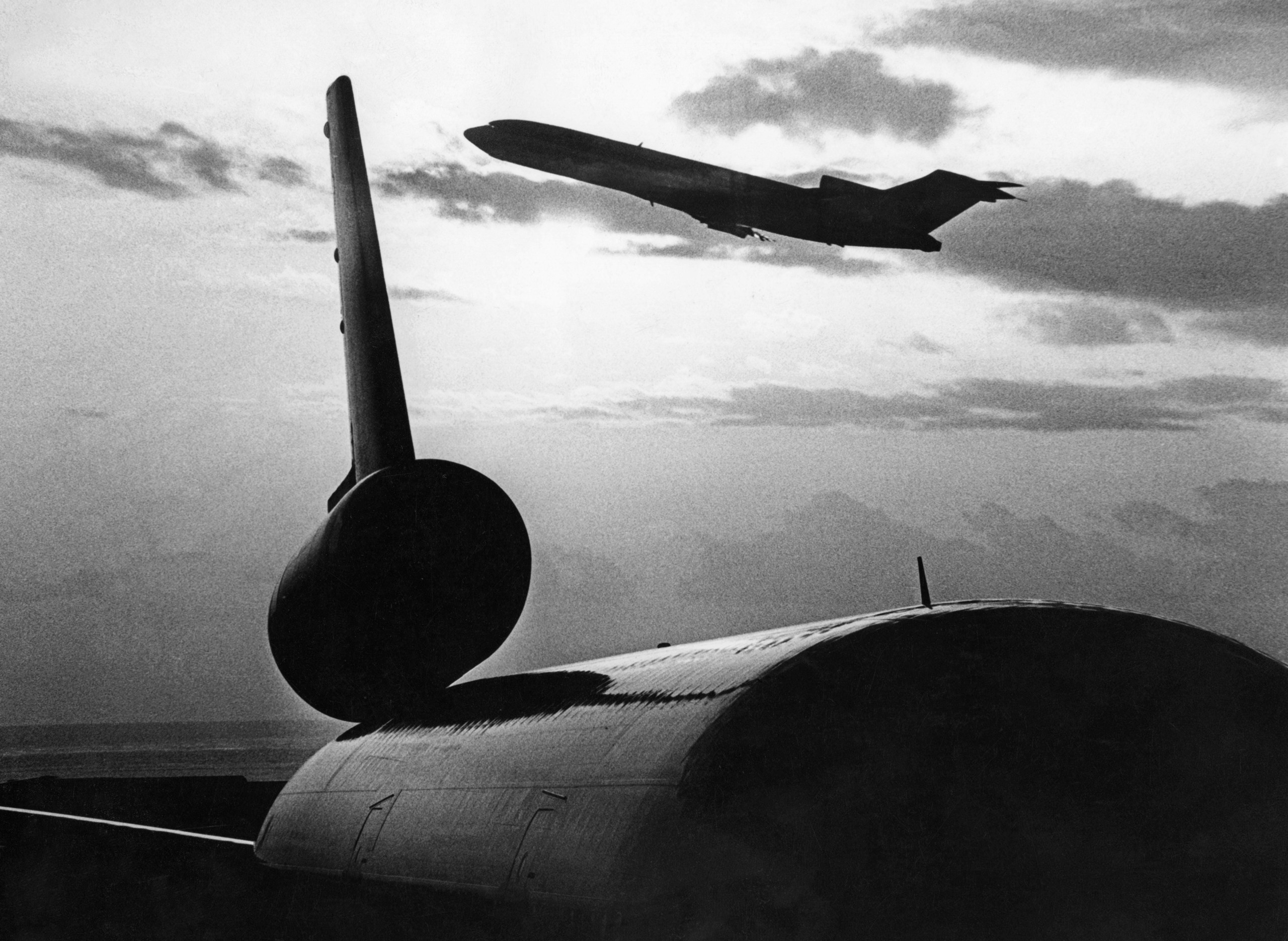 A high-contrast black and white photograph, likely from 1979, showing a Boeing 727 airplane flying high in the distance, silhouetted against a cloudy sky. The foreground is dominated by the large, dark silhouette of an aircraft tail section lying on the ground, creating a stark visual contrast.