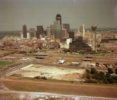An aerial photograph of the **Dallas, Texas skyline** taken in 1984, showing a dense cluster of downtown skyscrapers, including the tall, cylindrical Reunion Tower on the right. In the foreground are large, undeveloped dirt lots, elevated highways, and roads, illustrating the city's infrastructure and ongoing urban development.