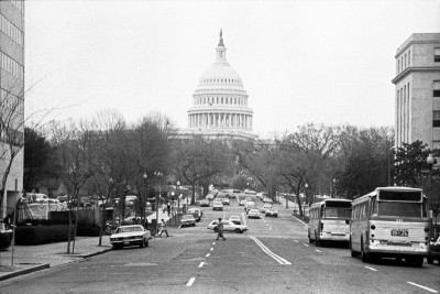 A black and white photograph from 1976 of a street scene in Washington, D.C., looking toward the U.S. Capitol Building dome in the distance. The wide street is lined with bare trees, cars from the 1970s, and two buses parked on the right, with several pedestrians crossing in the middle ground.